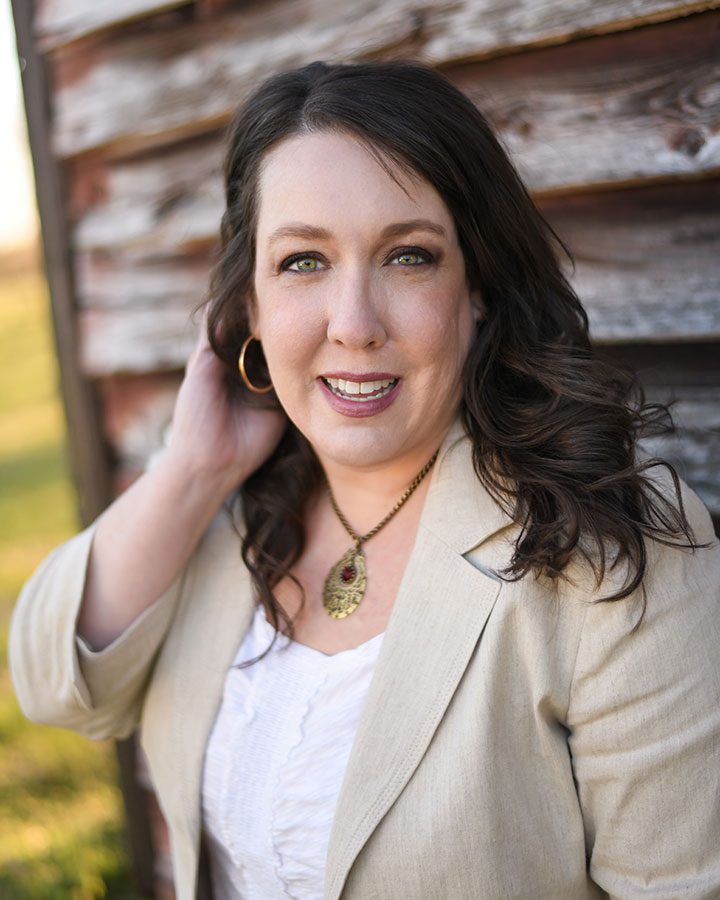 Woman with long brown hair, wearing a beige blazer, white blouse, gold necklace, and hoop earring, standing outdoors next to a weathered wooden wall.